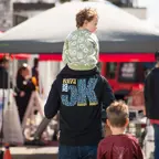 Dad with a 5K T-shirt on with a children on his shoulders walking in the Expo