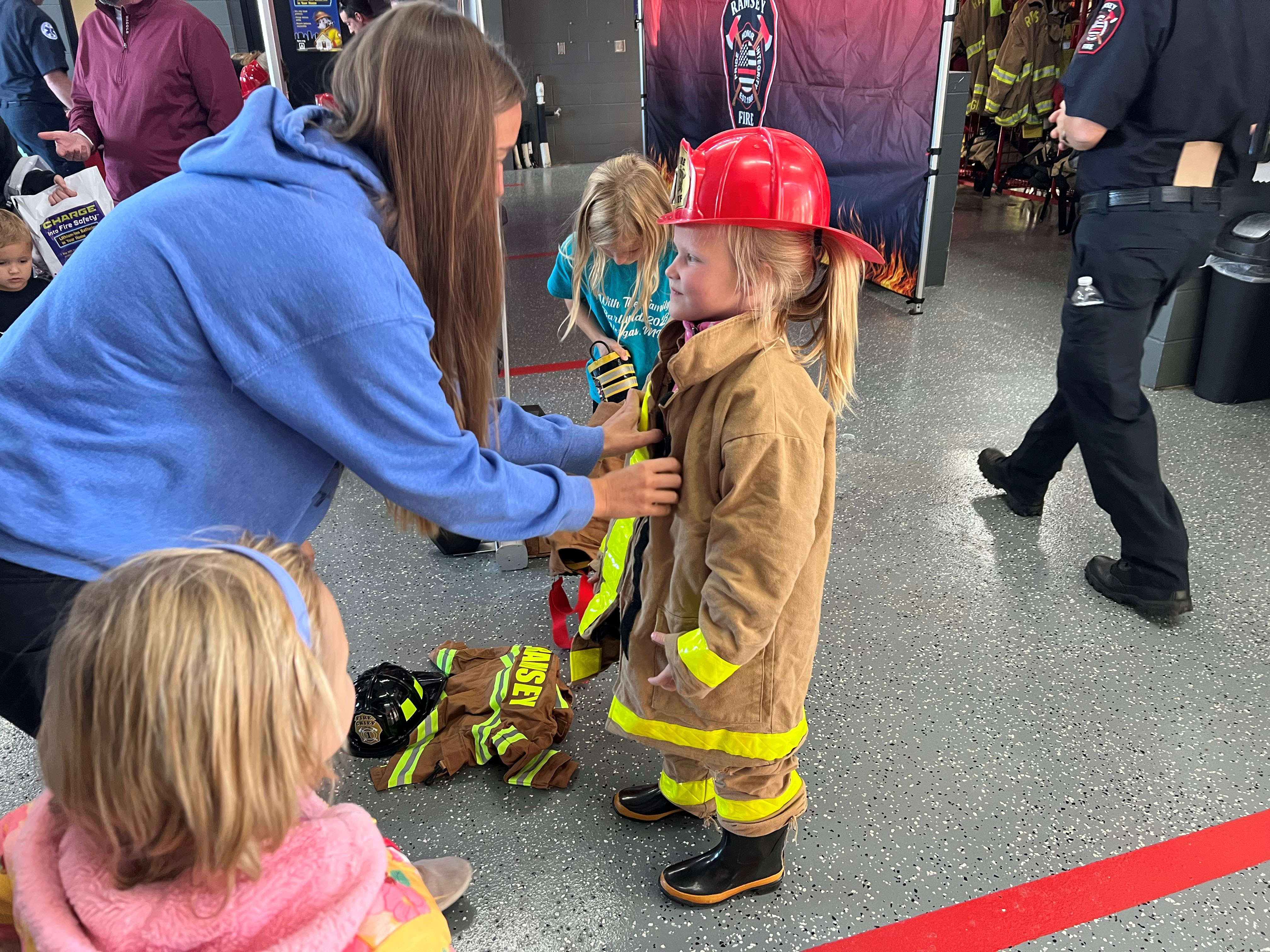 A mom is helping a little girl dress in firefighting attire, jacket, pants, boots and helmet