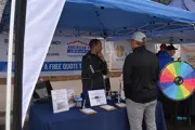Two Caucasian men in there 40s talking under a canopy with a table between them spin the wheel right