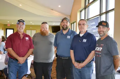ive adult men stand side by side indoors in a banquet-style room with beige walls, large windows, white drapes, round tables covered in white tablecloths, and a patterned carpet. A blue banner in the background reads in part “Economic Development Authority EDA” and “RAMSEY.”