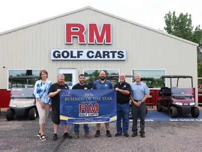 Six people pose in front of RM Golf Carts in Ramsey, Minnesota. They are holding a banner for the 2026 Business of the Year Award, presented by the Ramsey Economic Development Authority.