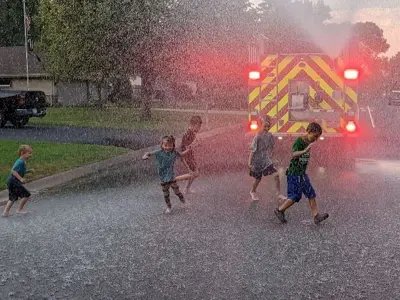 Fire truck in the middle of a residential street, 5 small children playing a water spray 