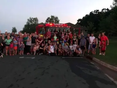 Approximately 50 people posing in 4 rows in front on a fire truck on a hot summer day