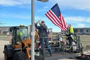 Two male workers in a basket on a front end loader in air attaching short flag poles to light poles