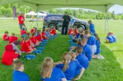 two rows of kids sitting in the grass under a canopy police officer instructing CPR