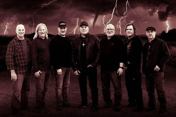 Seven male band members standing in a line, with storm clouds in the background