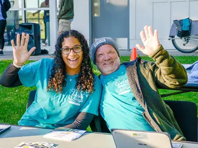 A female and male volunteer sitting a table waving to the camera