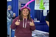 Woman in hoodie giving double thumbs up, wearing a pink balloon hat at a vendor booth.
