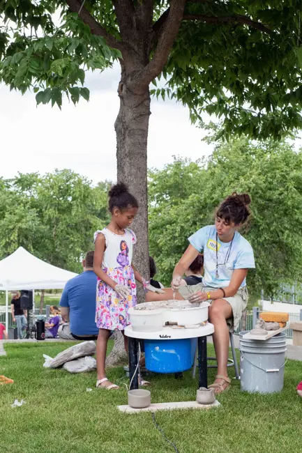 Outside in a park, a woman sitting at a potters wheel with a little girl watching her