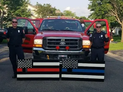 Two fire fighters posing in front of a rescue truck with doors open 2 wooden signs in front