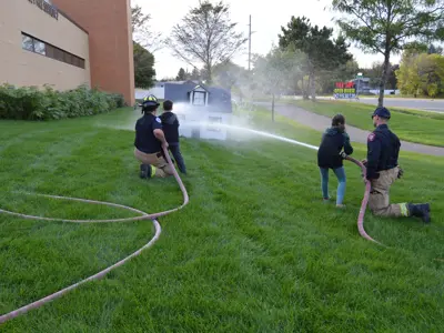 Two children being helped by two firefighters spray a wooden house with water from two fire hoses