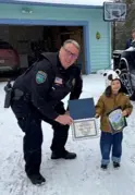 An officer crouched down next to a small child holding a certificate outside in the snow. 