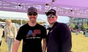 Two Caucasian men in their 30s in baseball caps and sunglasses standing beside each other smiling 