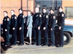 Eight Police Department staff from 1986 standing in a row outside in front of a police car. 