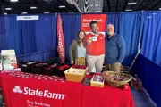Man & woman smiling at State Farm booth with a cardboard cutout of Jake from State Farm holding mug.